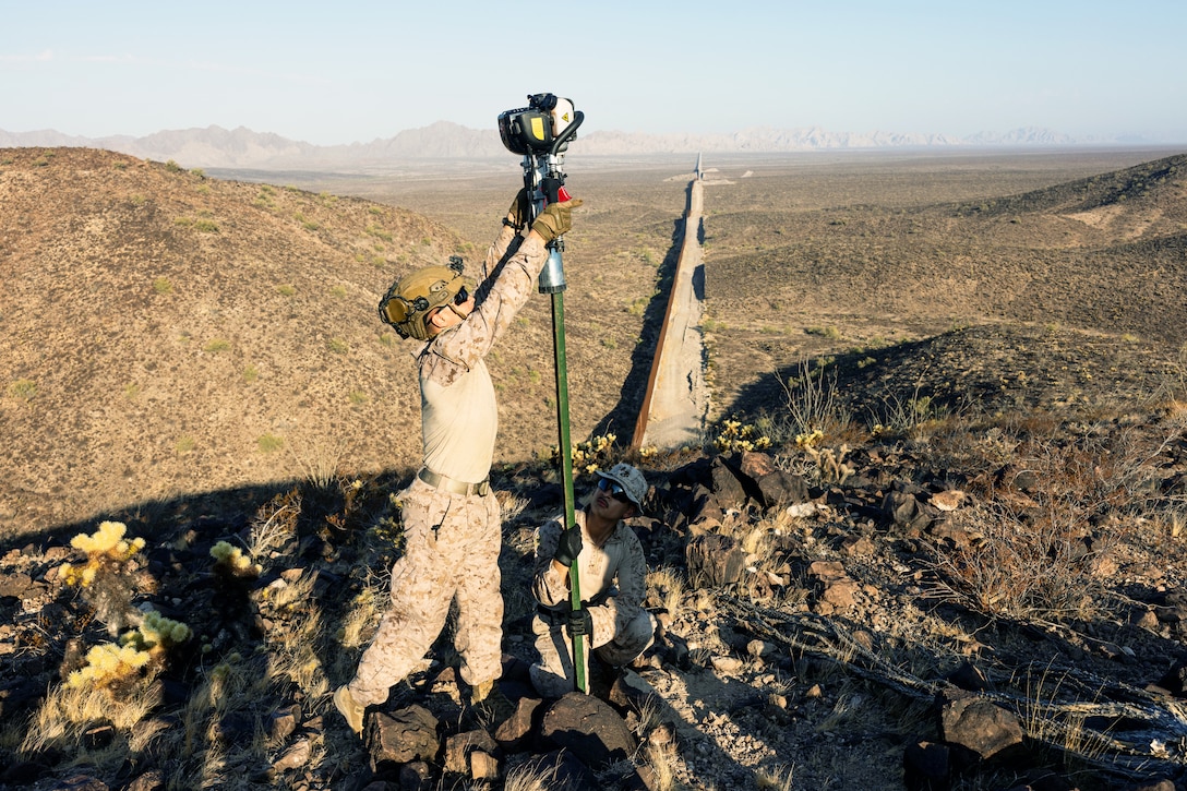 A Marine stands to secure a tall sign while another Marine crouches down to support on a desert-like hill on a sunny day.