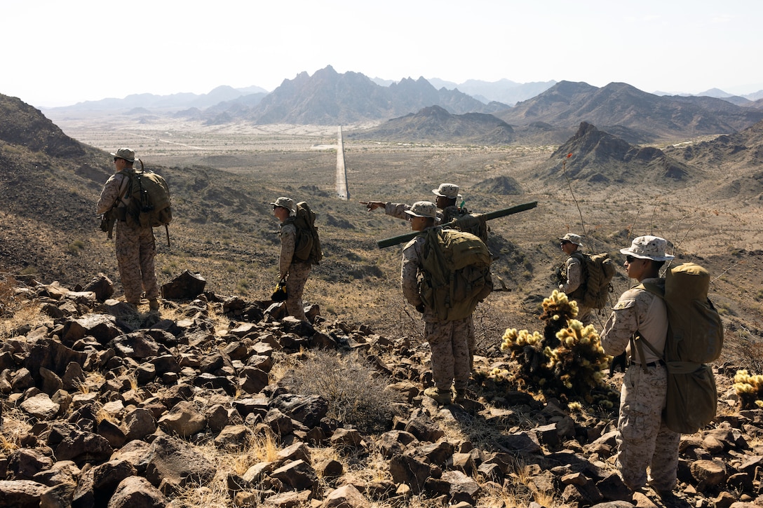 Marines with backpacks stand on a rocky ridge looking over a desert-like valley with mountains in the distance on a sunny day.