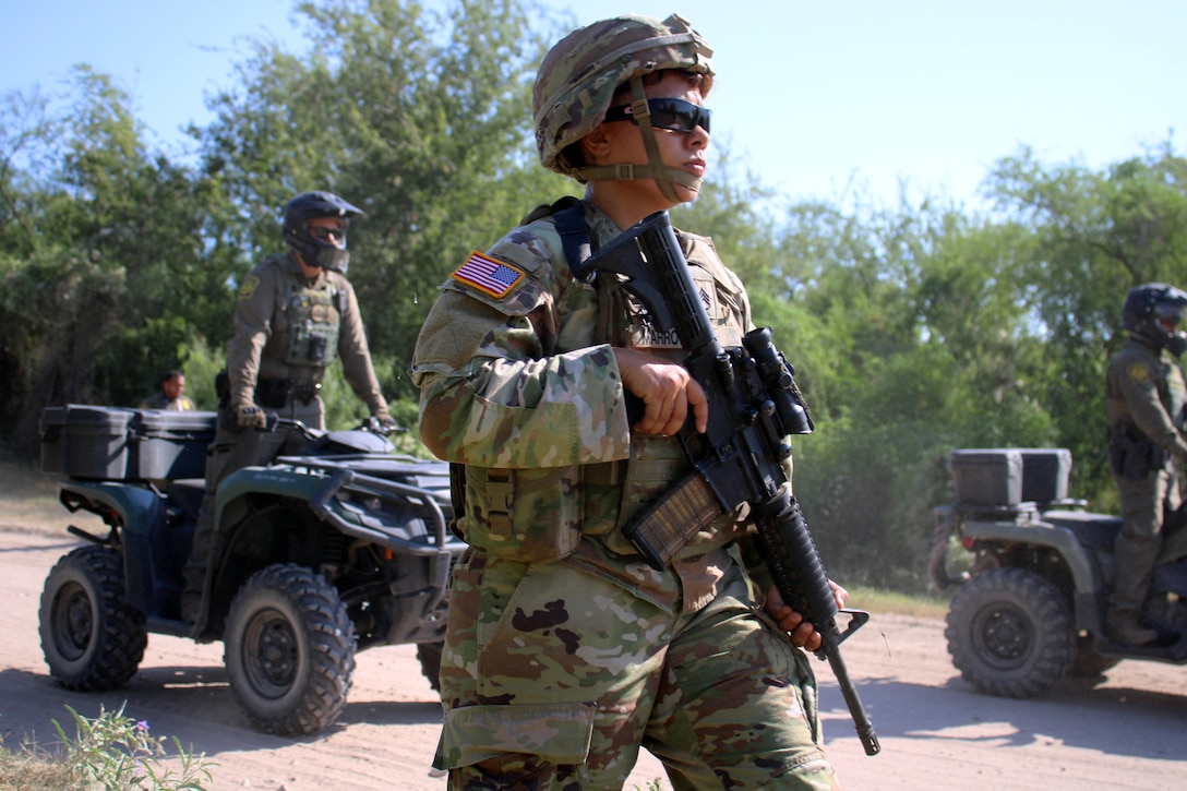 A soldier holding a service weapon walks next to two other soldiers on quads outside on a dirt path with trees behind.