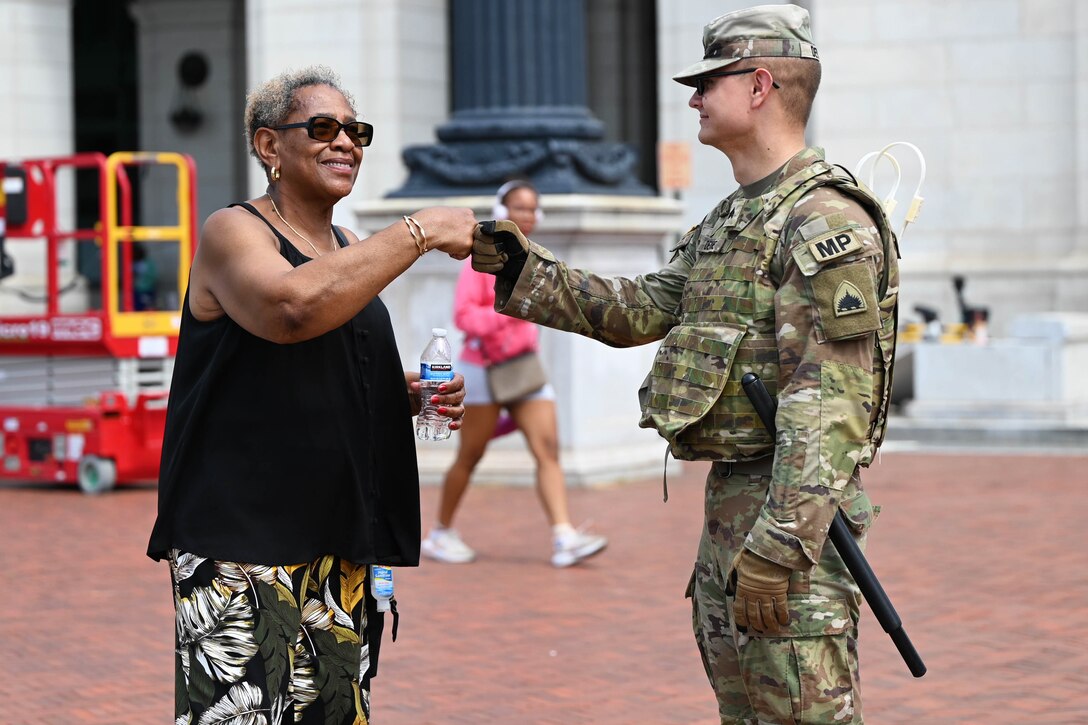 A guardsmen fist-bumps a person wearing civilian clothes on an outdoor brick patio, with another person and a building in the background during daytime.