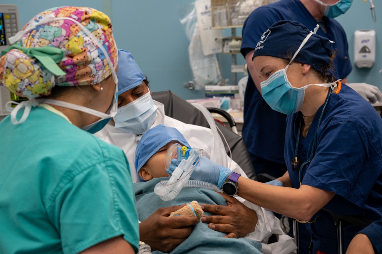 A sailor in blue medical scrubs places an oxygen mask over the face of a child who is lying in the arms of a person in medical scrubs. Two other people in medical scrubs are around the child.