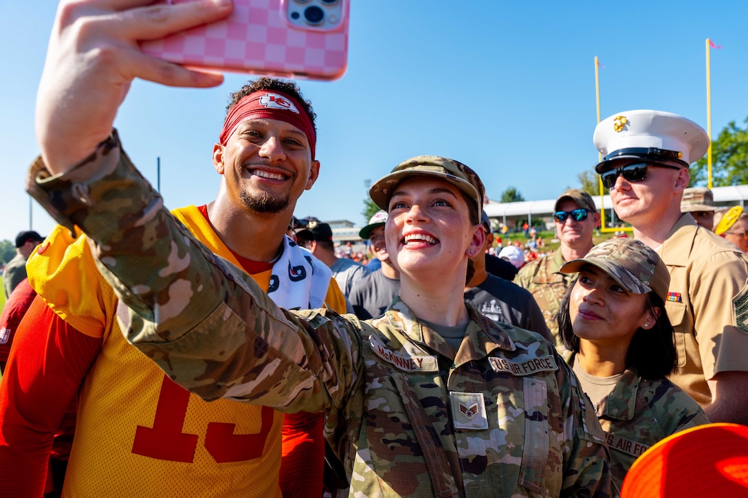 An airman smiles while taking a selfie with an NFL quarterback on a crowded football field on a sunny day.
