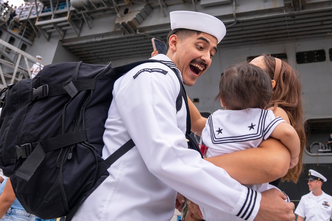 An excited sailor hugs a person carrying a baby in front of a ship.