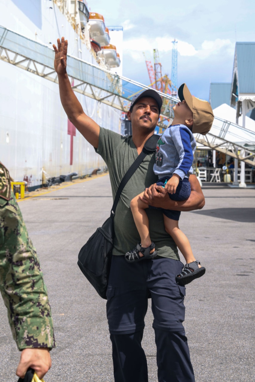 A person in casual attire waves while carrying a child. The child is wearing a hat and looking up at a large ship that appears in the background.