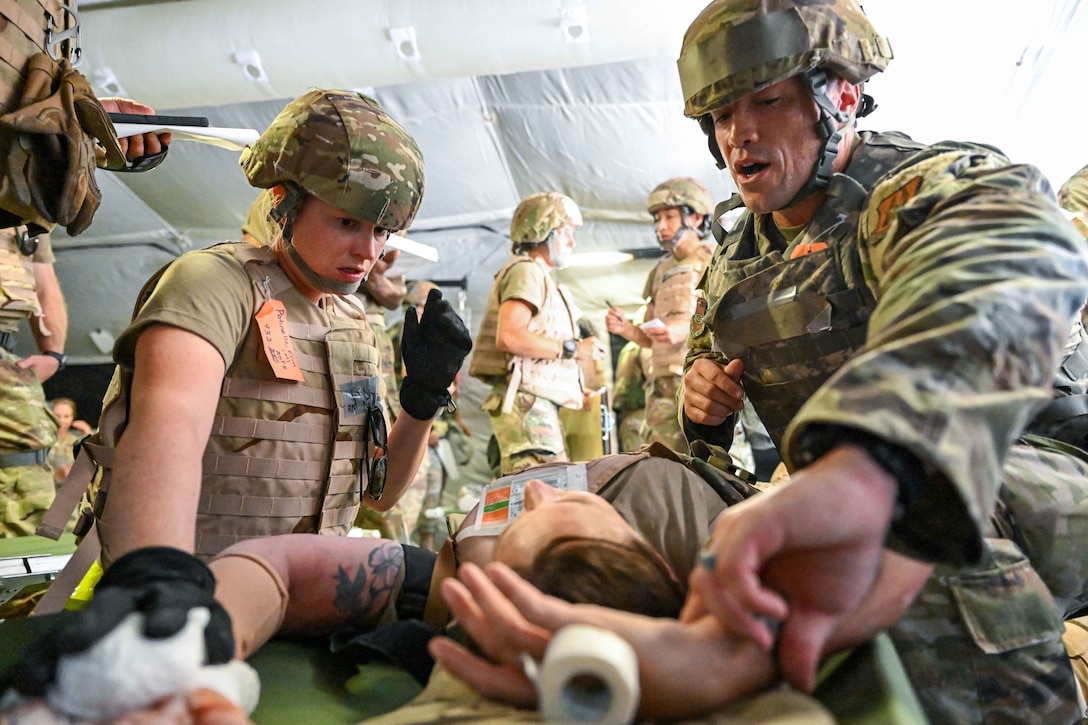 Two airmen provide medical care to a dummy on a gurney in an inflatable tent as fellow service members talk in the background.