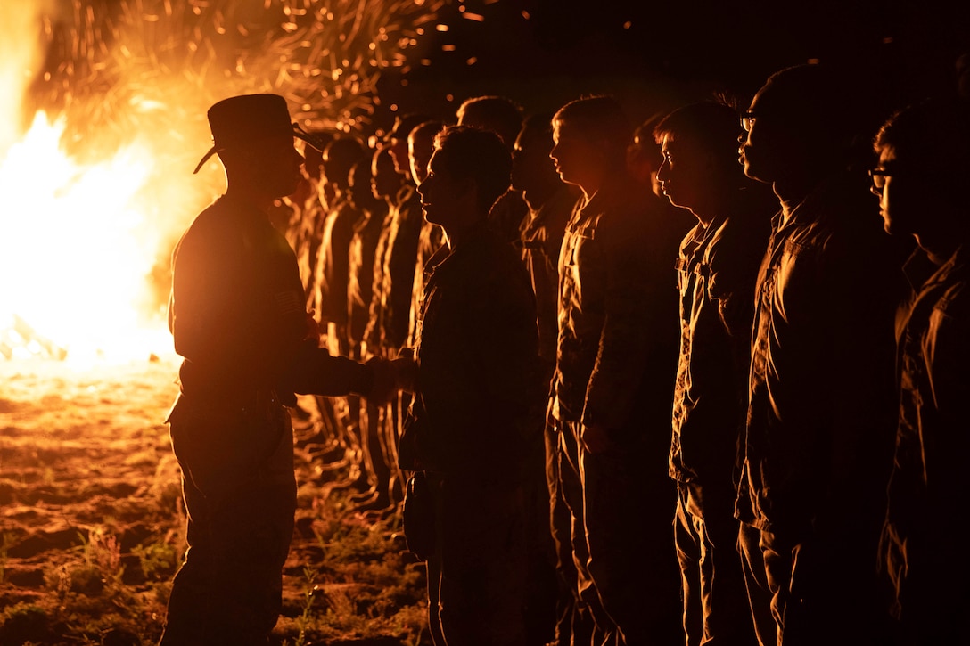 A command sergeant shakes hands with a soldier standing in formation with fellow soldiers on the sand in the dark as a bonfire creates large flames to the left.