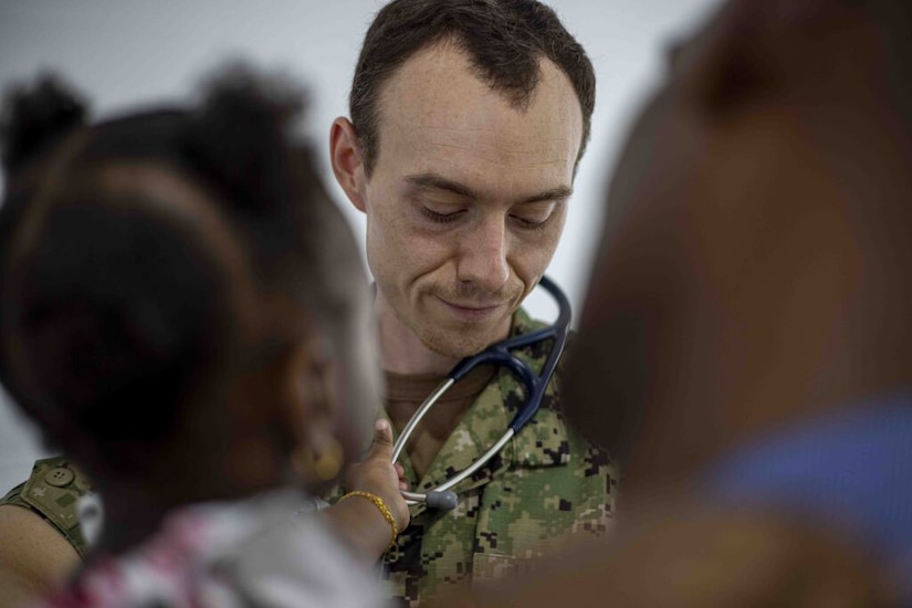 A blurry image of a child in the foreground grabs a stethoscope that is around the neck of a sailor in a camouflage uniform.