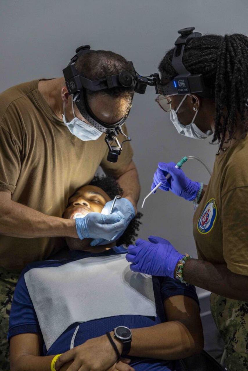 A sailor wearing a brown T-shirt and medical gear places a dental instrument in the mouth of a person in casual attire who is lying in a chair. A sailor wearing a brown T-shirt and medical gear is holding a dental instrument and standing to the right of the person lying in the chair.
