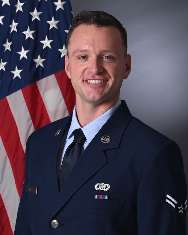 An official photo of A1C Christopher Hall standing in front of the American flag wearing the service dress uniform.