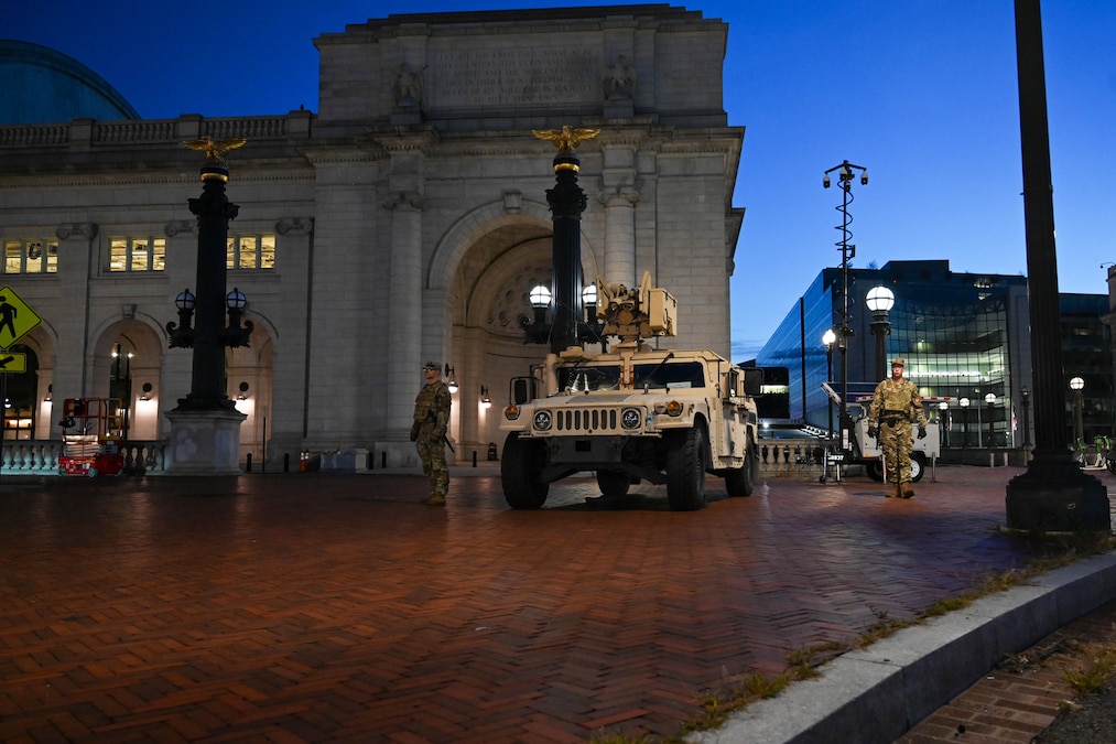 Two guardsmen walk near a military vehicle parked near city building under a deep blue sky.