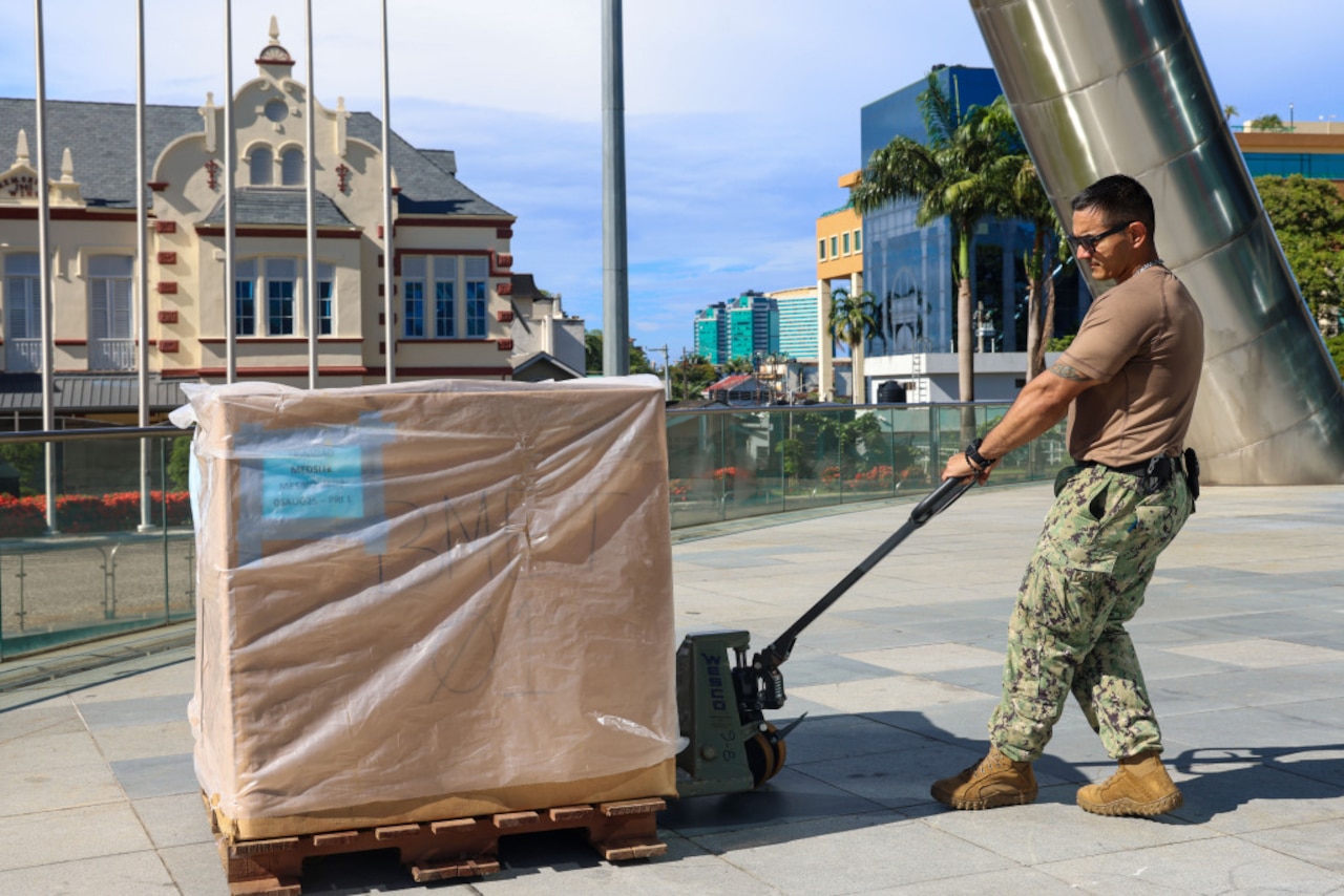 A sailor in a brown T-shirt and camouflage pants moves a large box using a pallet jack.