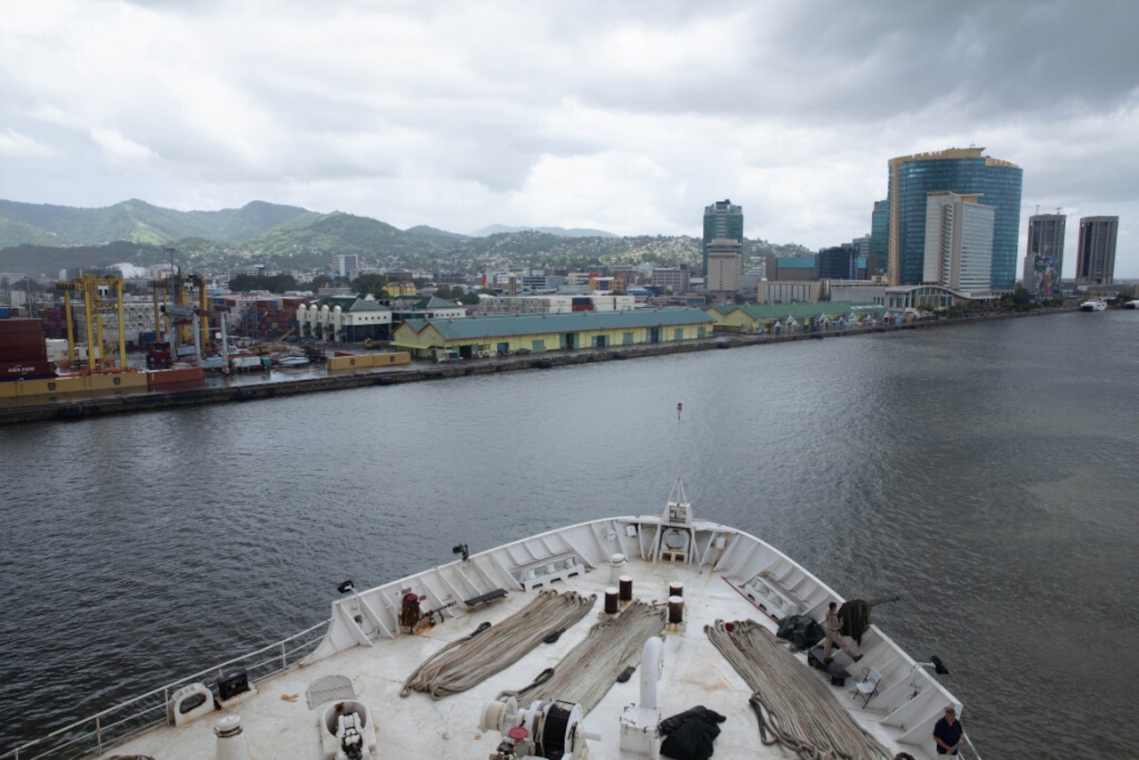 A large white ship is sailing in the foreground with buildings and mountains lining the background.