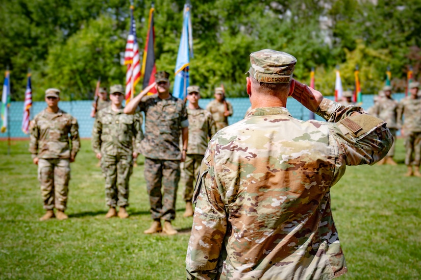 A man wearing a camouflage military uniform stands with his back to the camera and salutes a group of people also wearing camouflage military uniforms. Some are holding flags.