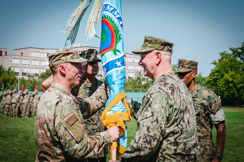 Two men wearing camouflage military uniforms smile as they both grasp a blue flag with yellow fringe. Other people in camouflage uniforms are in the background.