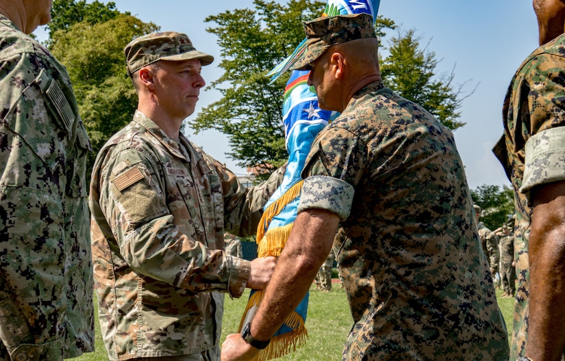 Two men wearing camouflage military uniforms smile as they both grasp a blue flag with yellow fringe. Other people in camouflage uniforms are in the background.