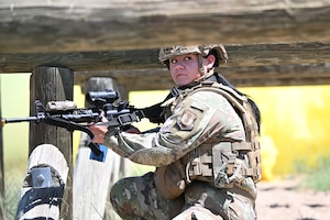 An airman in combat gear and a gun squats under a wooden obstacle
