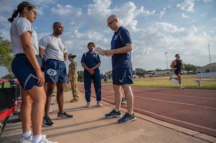 PT Trainer has his clipboard and is talking to a group of Airman who are about to take their PT test. Photo is outside on running track.