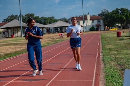 Trainer runs on the track in front of Airman coaching her and encouraging her to finish her run.