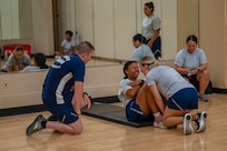 Airman doing pushups while observer is kneeling by. Another Airman is holding feet of Airman doing pushups. They are inside the gym.