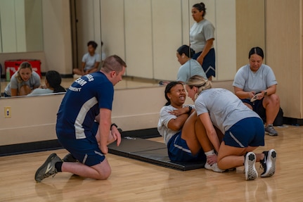 Airman doing pushups while observer is kneeling by. Another Airman is holding feet of Airman doing pushups. They are inside the gym.