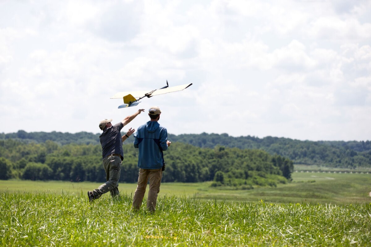 Two individuals wearing casual attire launch a small aircraft from a field into the air.