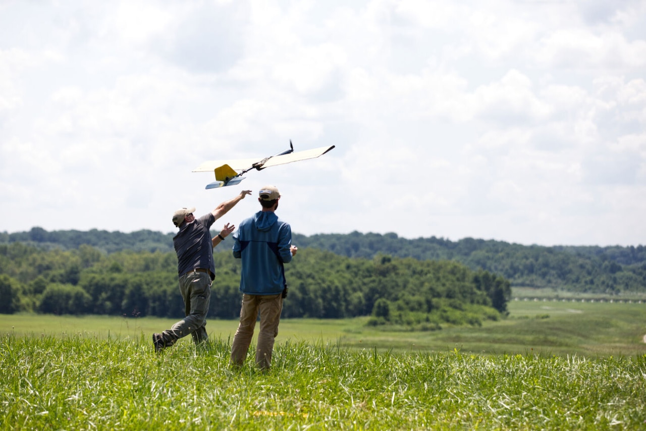 Two individuals wearing casual attire launch a small aircraft from a field into the air.