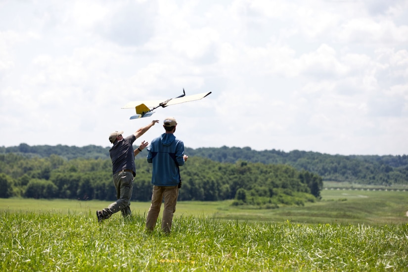 Two individuals wearing casual attire launch a small aircraft from a field into the air.