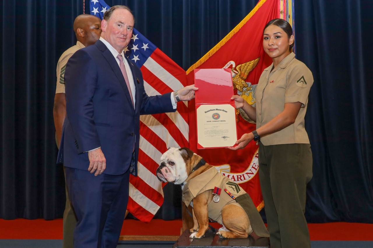 A dog, wearing a uniform with a military rank and a medal, sits on a platform. The dog is flanked by a man in a blue suit, and a woman in a military dress uniform. The man and woman are holding a certificate.