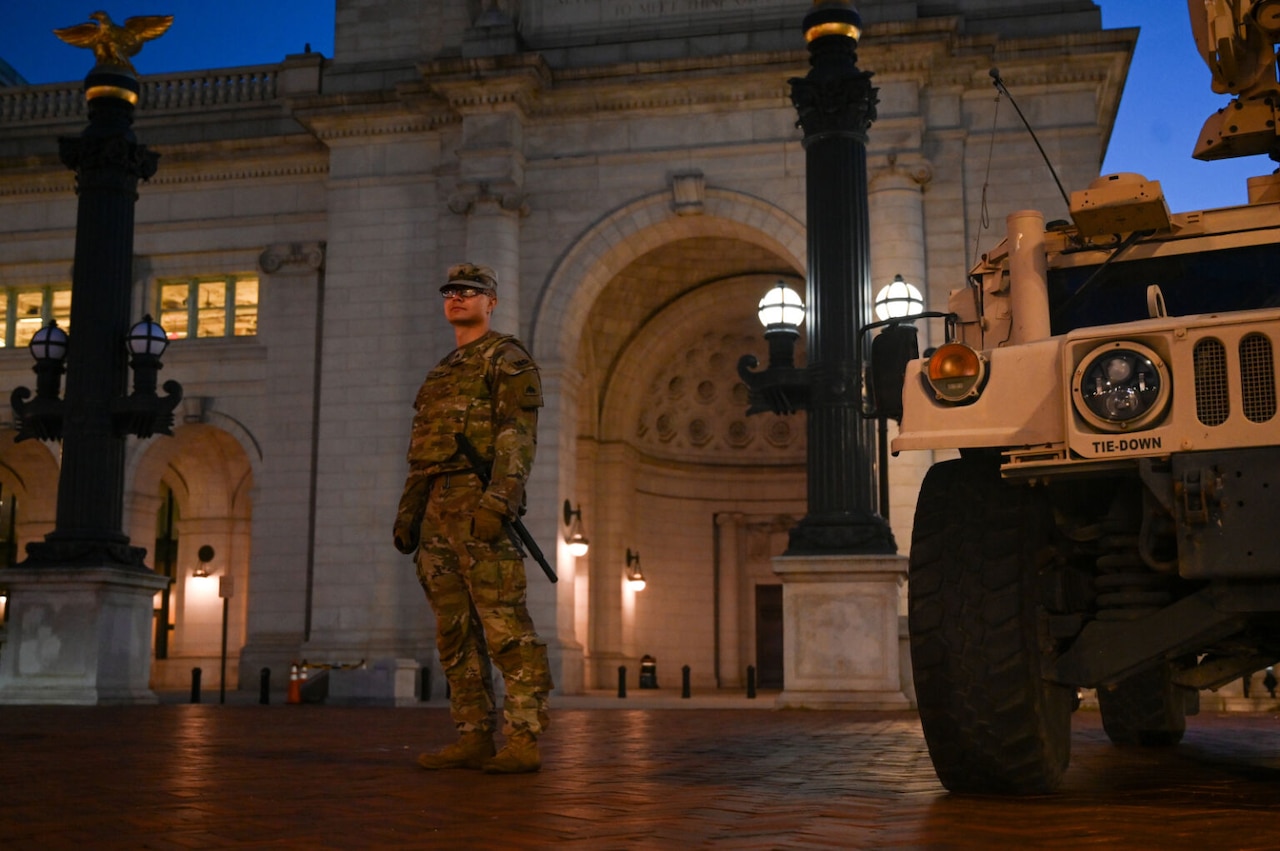A uniformed soldier stands alone near a military vehicle. Behind the soldier is a large building with an arch and a vault.