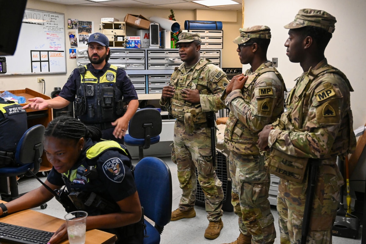 In a room, three uniformed military personnel with the letters "MP" on their sleeves talk with a man in a police uniform. A woman wearing a police uniform, sits at a computer.