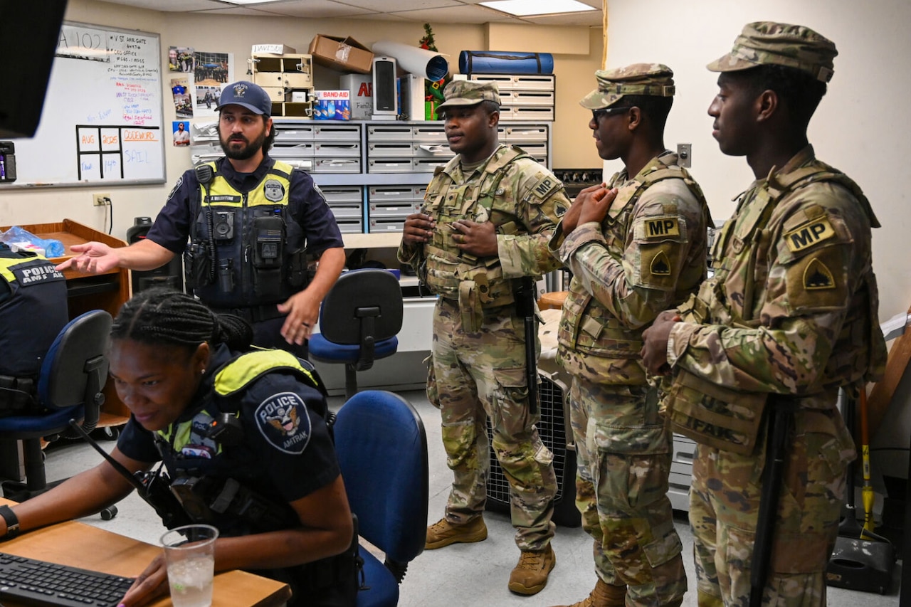 In a room, three uniformed military personnel with the letters "MP" on their sleeves talk with a man in a police uniform. A woman wearing a police uniform, sits at a computer.