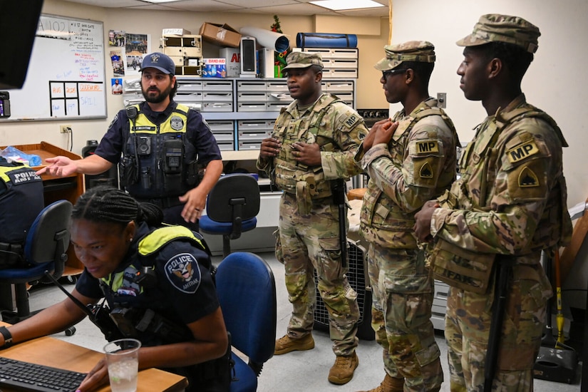 In a room, three uniformed military personnel with the letters "MP" on their sleeves talk with a man in a police uniform. A woman wearing a police uniform, sits at a computer.