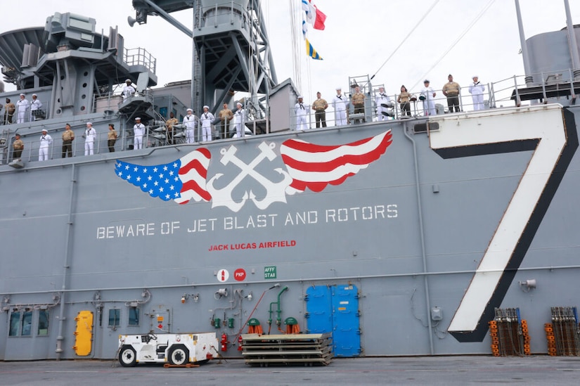 People wearing formal military uniforms stand along the rails of a ship while it is docked. The words, “Beware of jet blast and rotors” and “John Lucas Airfield” are written on the side of the ship, along with a giant number seven.