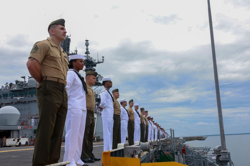 People dressed in formal military uniforms stand at attention along the rails of a ship.