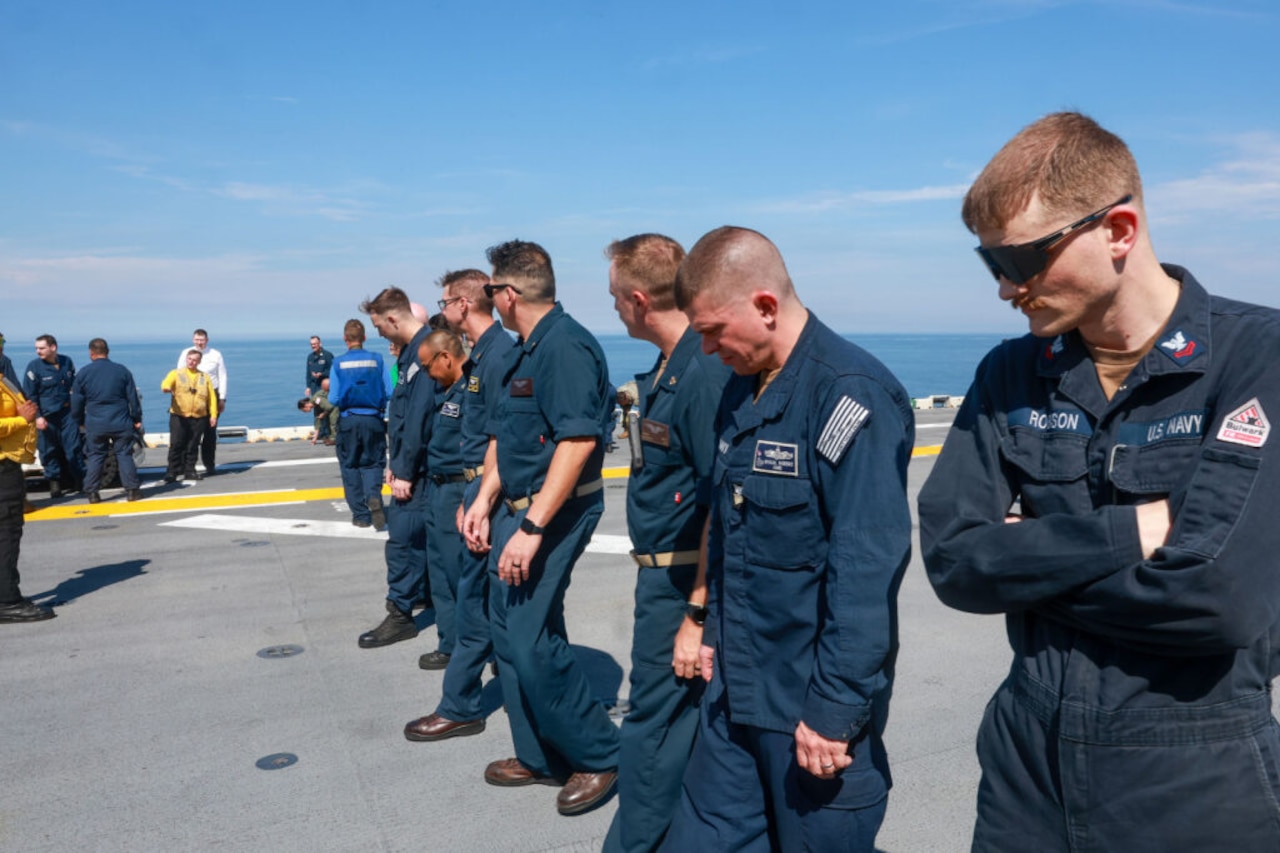People wearing blue military uniforms look down as they walk along the deck of a ship searching for debris.