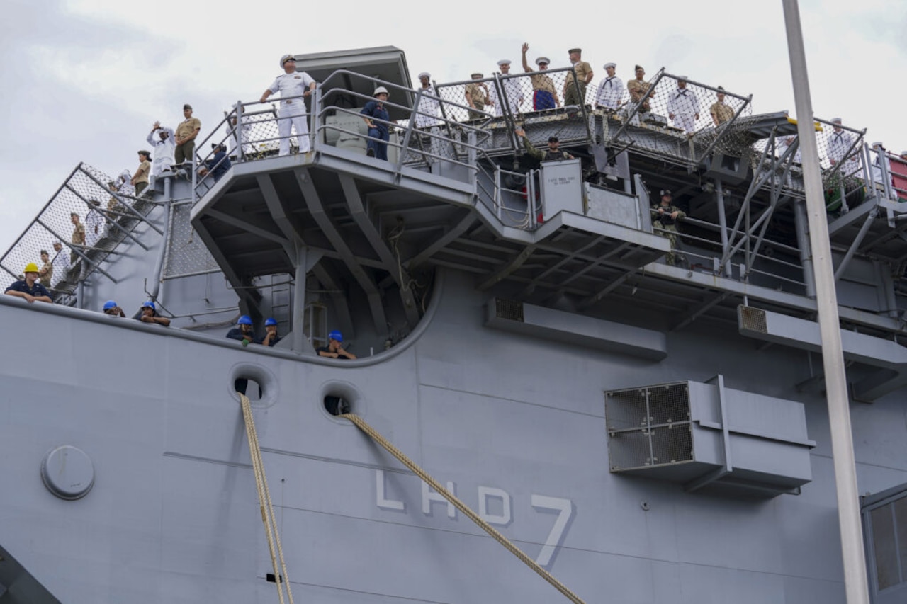 People wearing formal military uniforms stand along the rails of a ship as it heads out to sea, some of them waving.