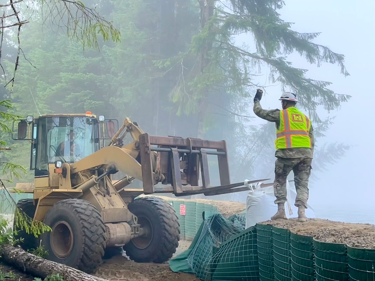 A person wearing an Army camouflage uniform, reflective vest and a helmet stands on a flood barrier and guides a forklift driven by another person.