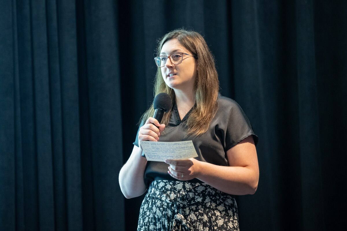 A woman with brown hair, wearing glasses, a black blouse, and black & white flower print skirt, holds a microphone in one hand and large notecard in the other. She is standing in front of a dark curtain