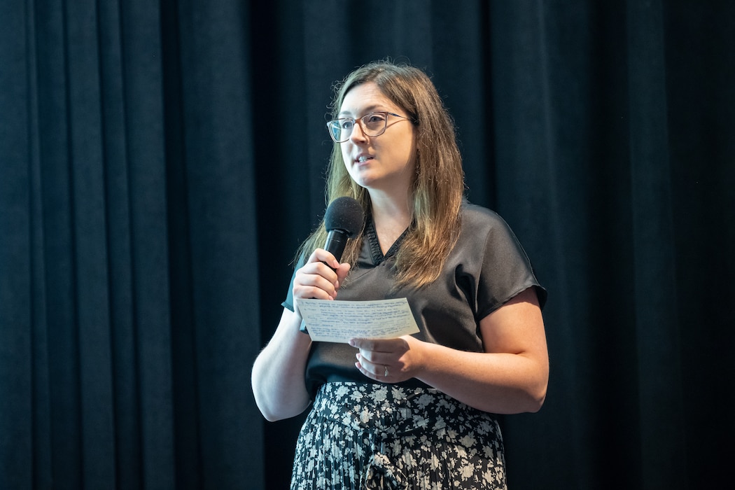 A woman with brown hair, wearing glasses, a black blouse, and black & white flower print skirt, holds a microphone in one hand and large notecard in the other. She is standing in front of a dark curtain