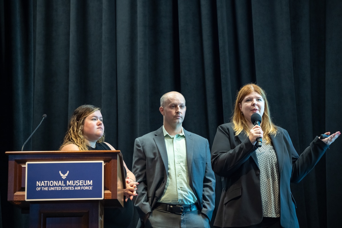 Two women and one man stand in front of a dark curtain. The woman on the left is standing behind a podium with a blue sign that reads National Museum of the United States Air Force in white text. The woman on the right holds a microphone and gestures with her hand