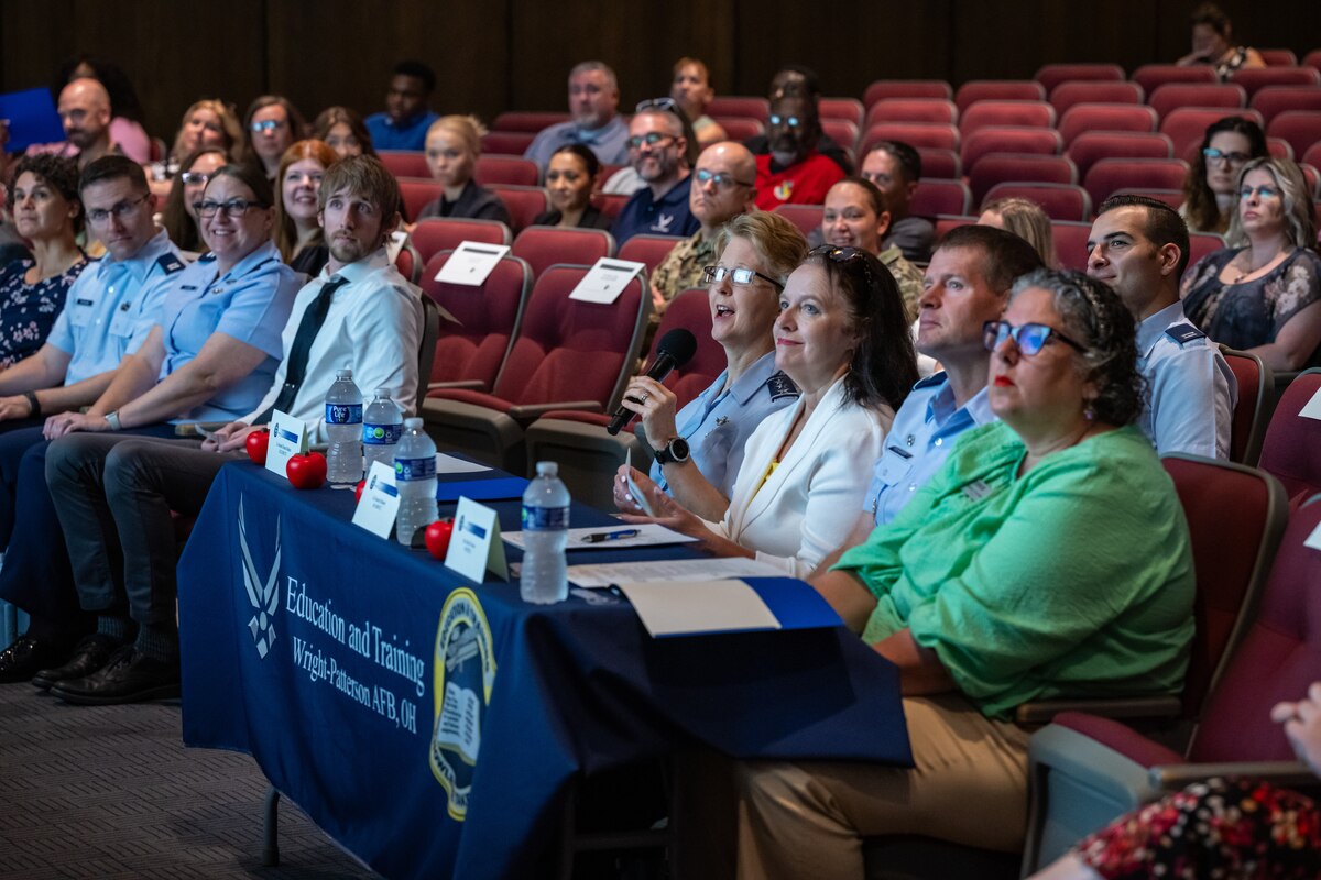 A mixture of civilian and military personnel scattered through auditorium seating. A group of four sit behind a small table with a blue tablecloth