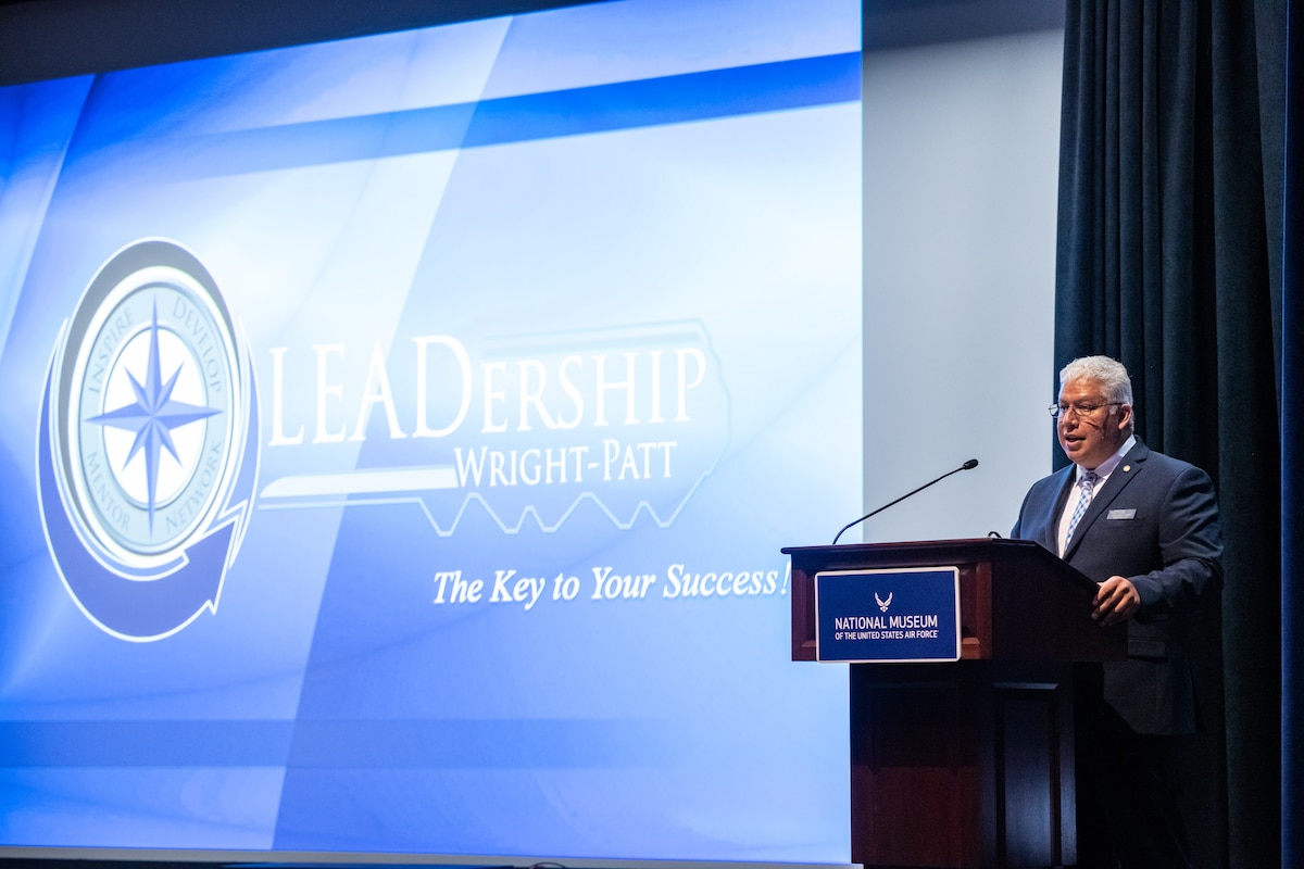 A white-haired man, wearing a dark suit, stands behind a podium in front of a large projection screen reading LEADership Wright-Patt.