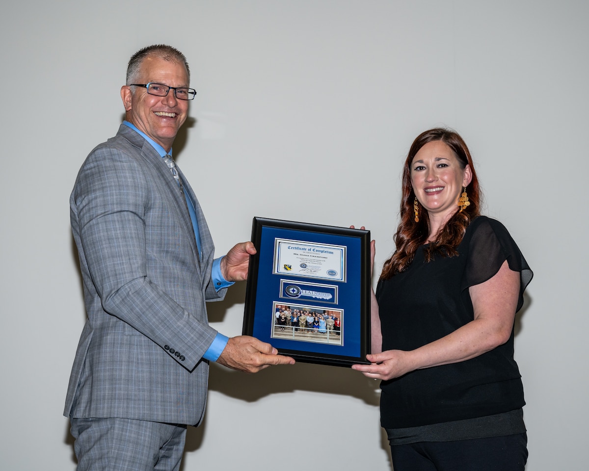 A tall man in a light grey suit presents a framed certificate to a woman in black blouse and pants