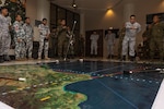 Uniformed military planners stand around a large map on the floor of a room to plan maneuvers.