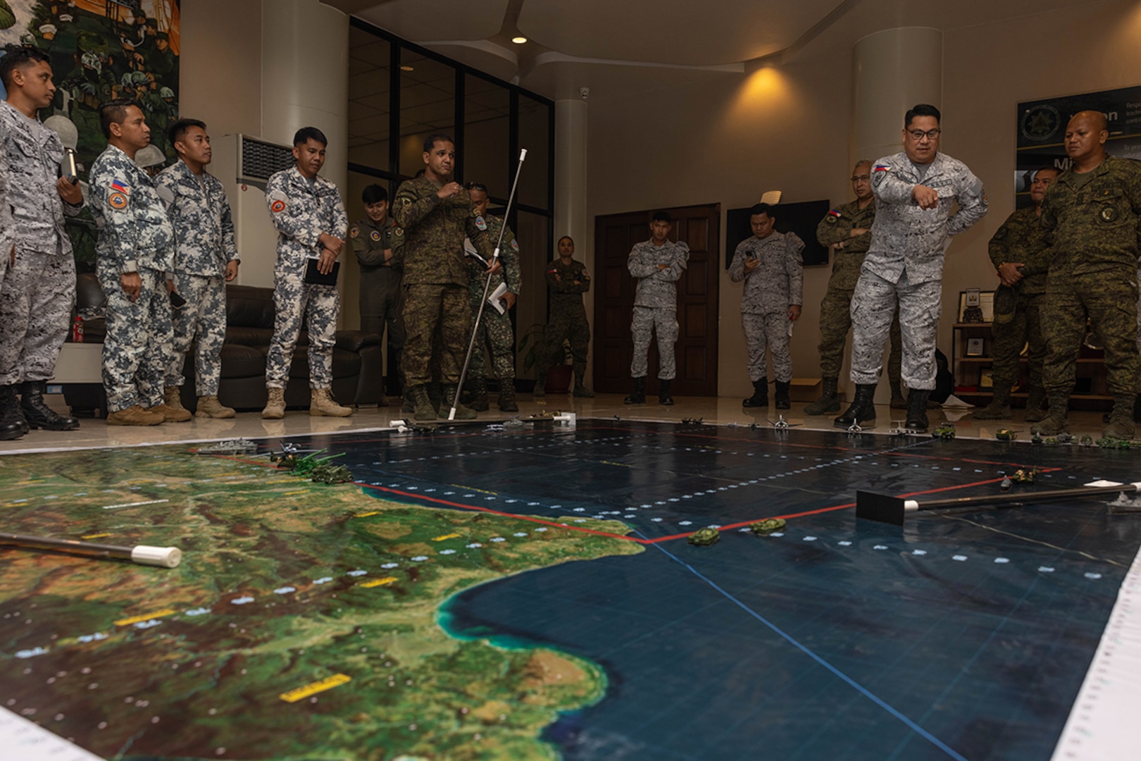 Uniformed military planners stand around a large map on the floor of a room to plan maneuvers.