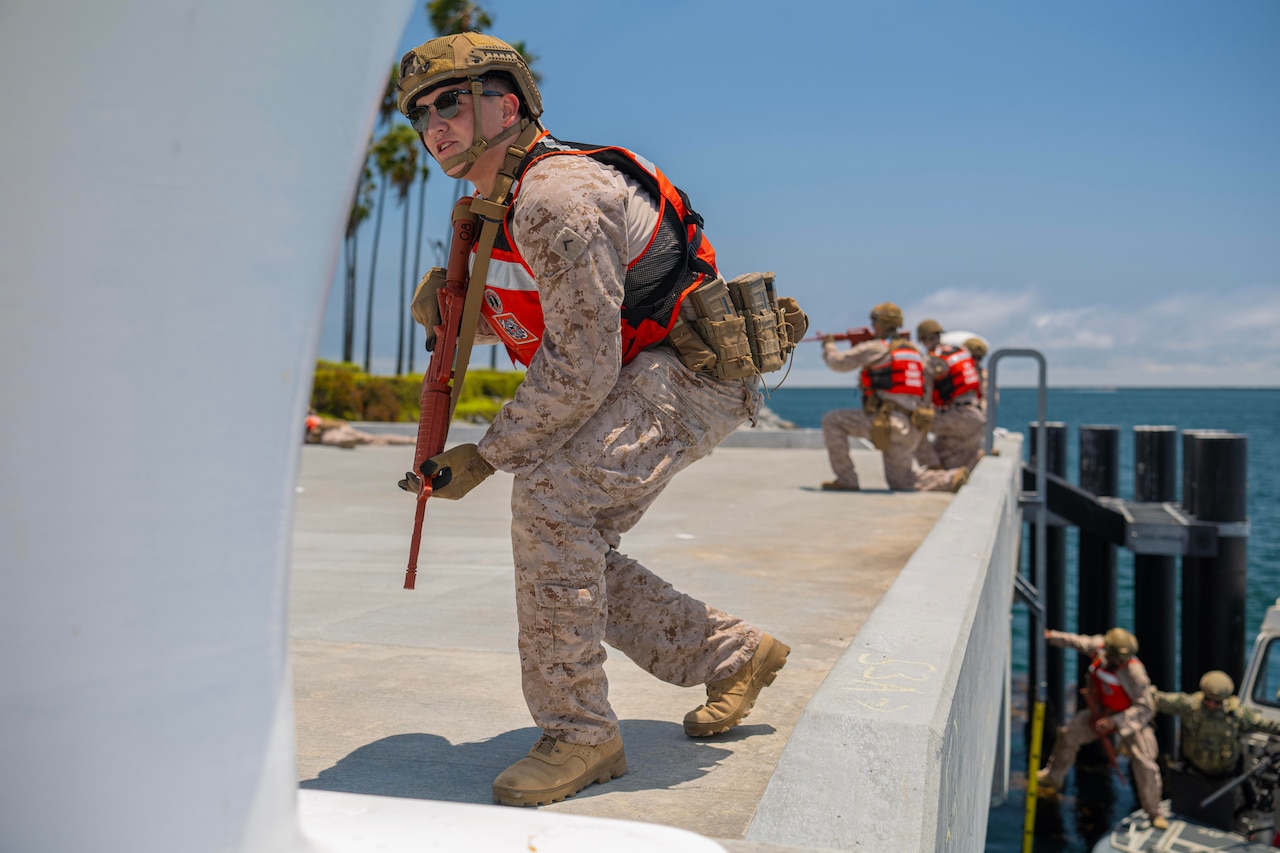 Marines in red vests and helmets hold service weapons on a concrete dock near the ocean after climbing up from a boat.