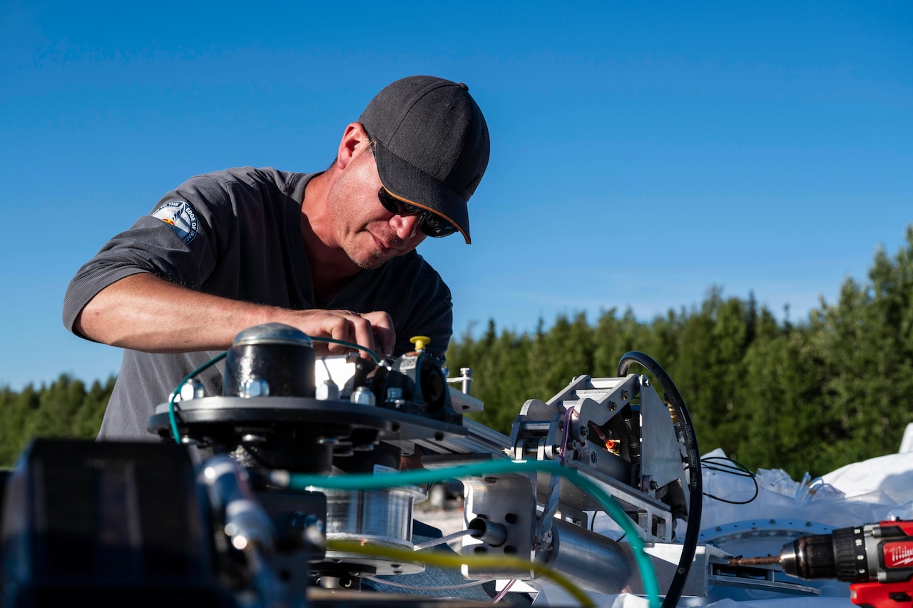 A person adjusts equipment for a high-altitude balloon outside under blue skies with a forest behind.