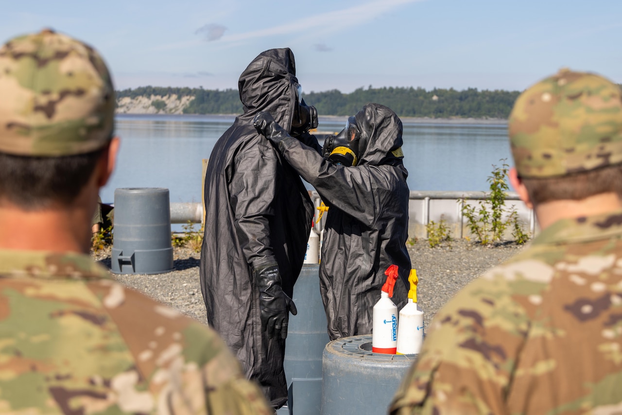 Two Marines in black hazmat suits stand in front of Coast Guardsmen near a harbor.