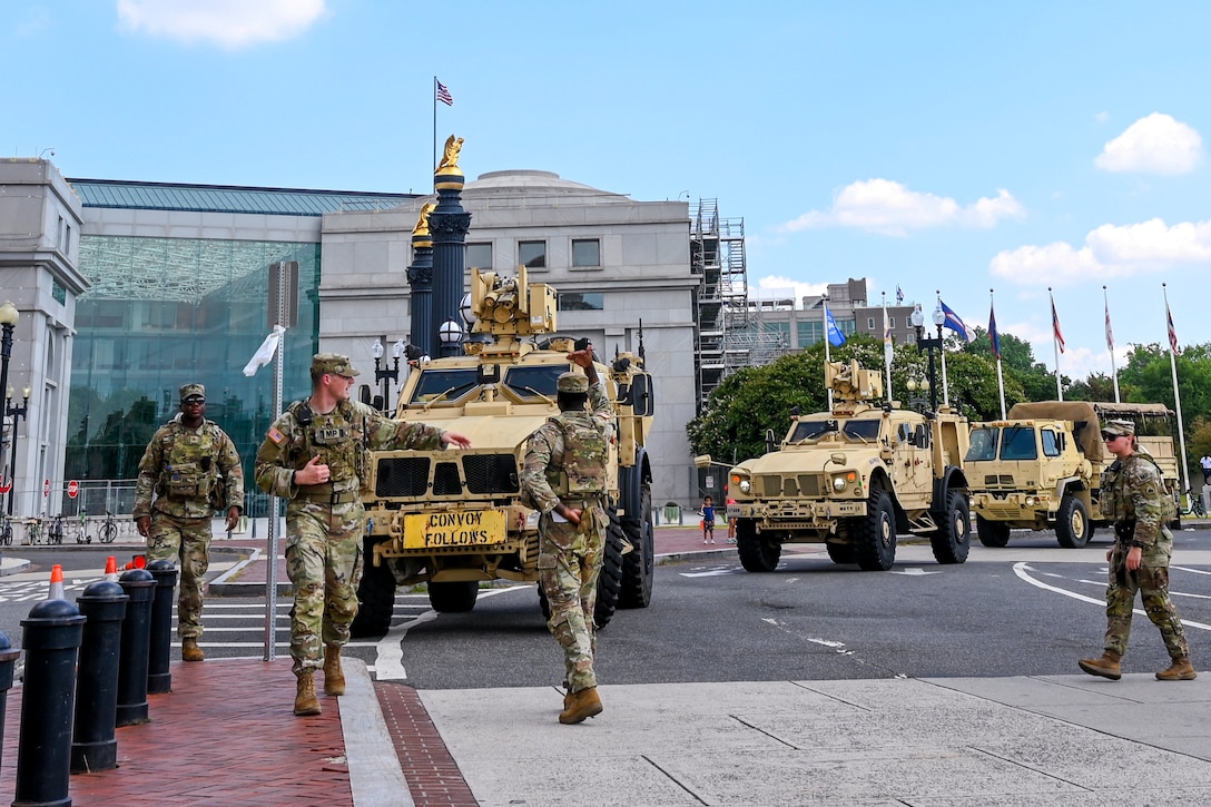 Guardsmen stand on a sidewalk and in a paved roundabout as three tactical vehicles pull to the side of the roundabout, with buildings, flags on tall poles and trees in the background on a sunny day.