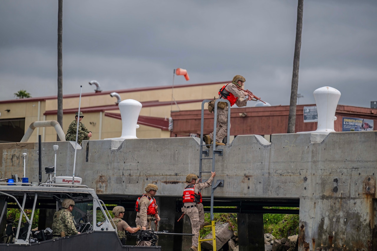 Marines in tactical gear climb up a ladder from a boat onto a concrete bridge under cloudy skies.